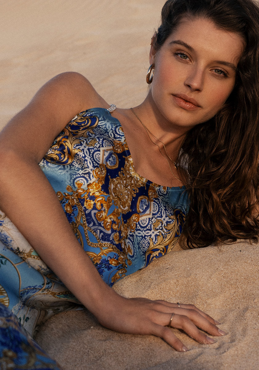 A woman with wavy brown hair and gold hoop earrings reclines on sand, wearing a blue and gold Angel-Print Seidenkleid by Princess Goes Hollywood. She looks softly at the camera, her left hand on the sand showcasing a simple ring in natural light.