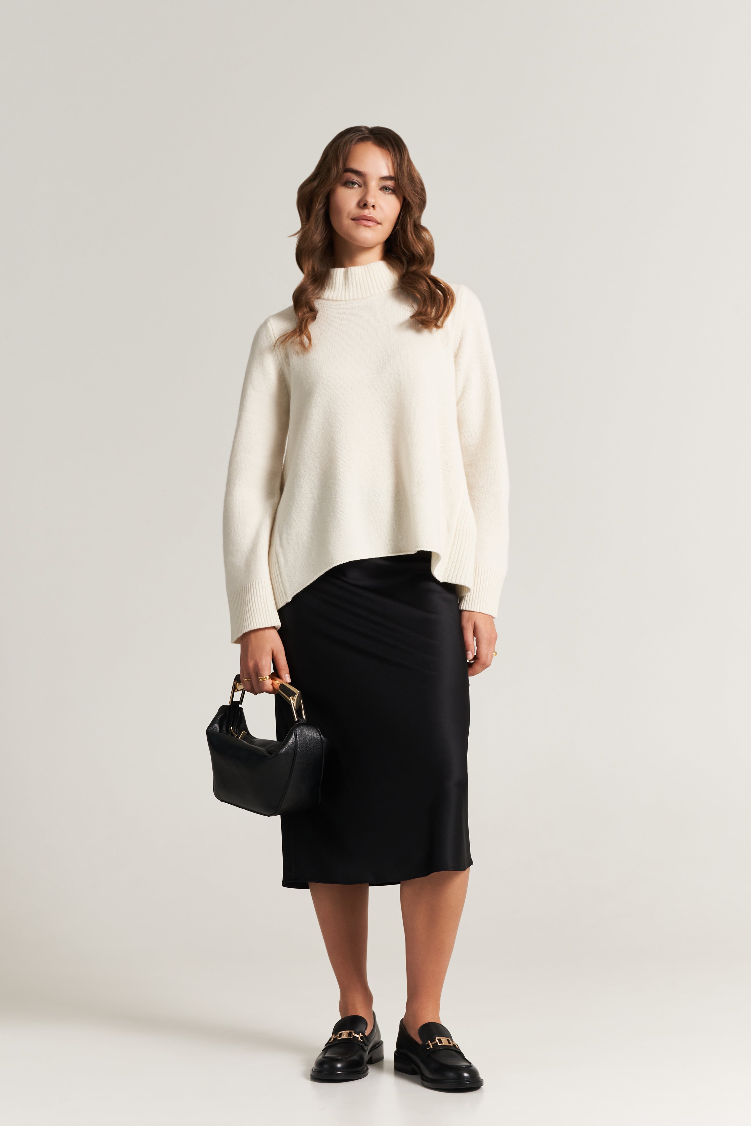 A woman stands against a plain light background. She wears a cream turtleneck sweater, a black midi skirt, and black loafers. She holds a black handbag with gold hardware in her left hand. Her brown hair is styled in loose waves.