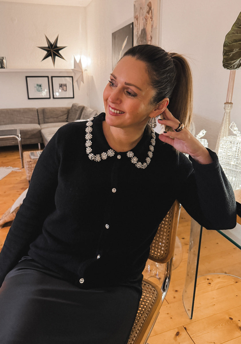 A woman with brown hair in a ponytail smiles indoors, wearing a Frogbox by Princess Strickjacke with a beaded floral collar. She sits on a chair in a warmly lit room with wooden floors, Kristalldeko, star-shaped decor, and wall art.