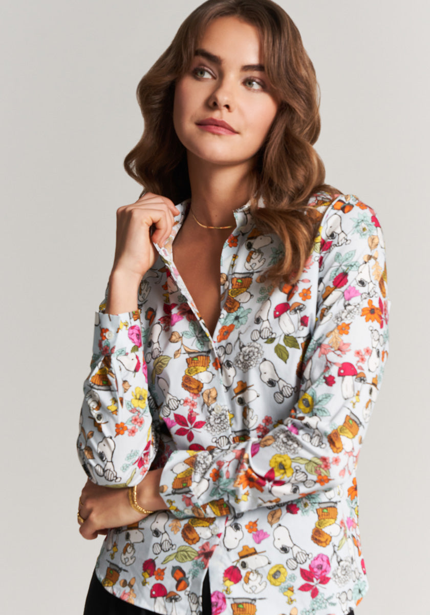 A woman with wavy brown hair wears the Frogbox by Princess Hemdbluse Fall Snoopy, a colorful blouse with cartoon animals and flowers. She stands against a plain background, holding her collar and gazing upward thoughtfully.