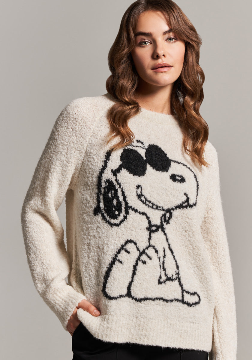 A woman with wavy brown hair wears a fuzzy cream-colored Frogbox by Princess Snoopy pullover featuring a large black outline of Snoopy in sunglasses. She stands confidently against a plain gray background.