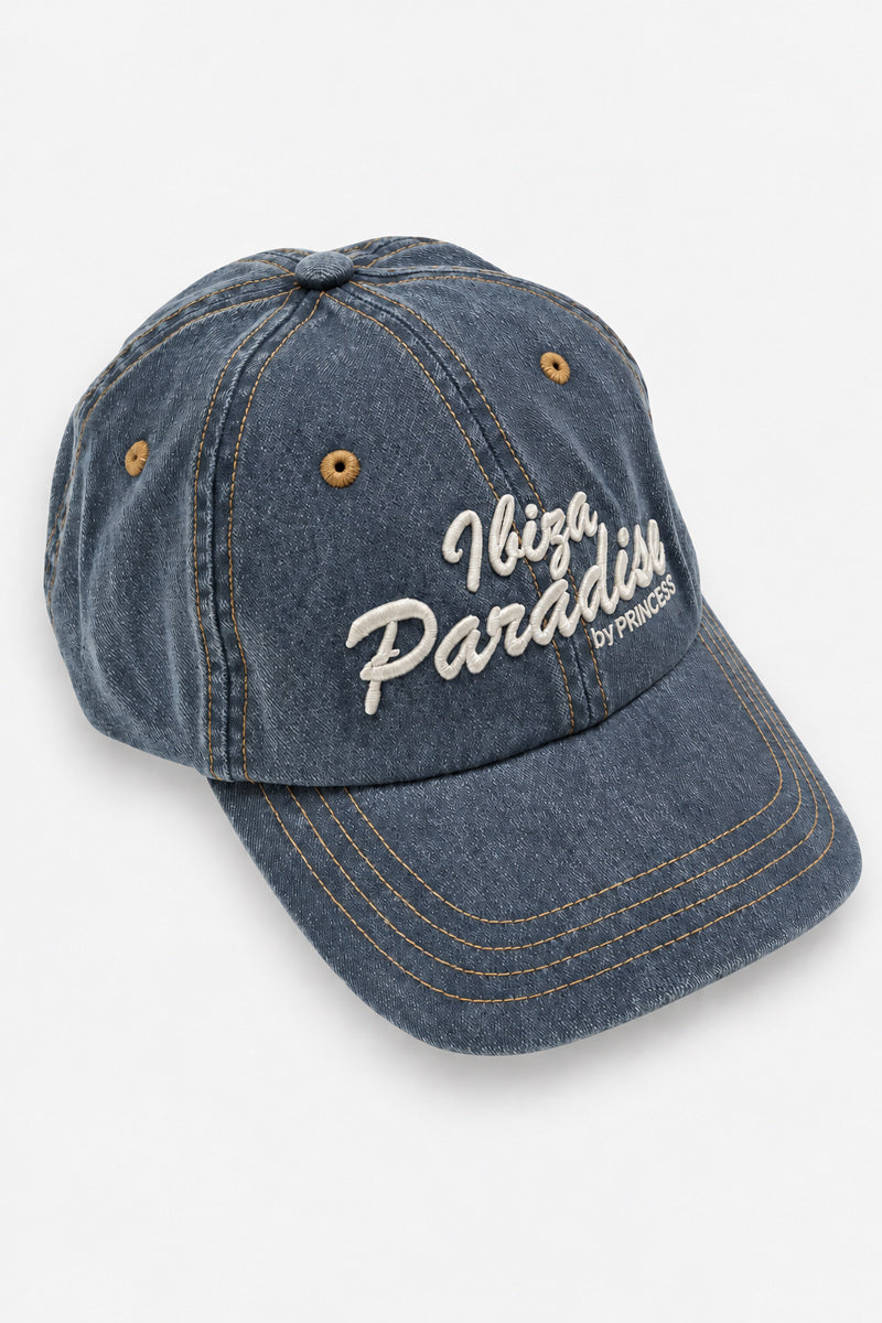 Auf weißem Hintergrund ist eine dunkelblaue Baseballkappe aus Baumwoll-Denim mit Acid-Wash-Effekt zu sehen. Die Ibiza Paradise-Kappe ist mit weißer Schrift bestickt und verfügt über kupferfarbene Ösen sowie goldene Nähte entlang der Krempe.