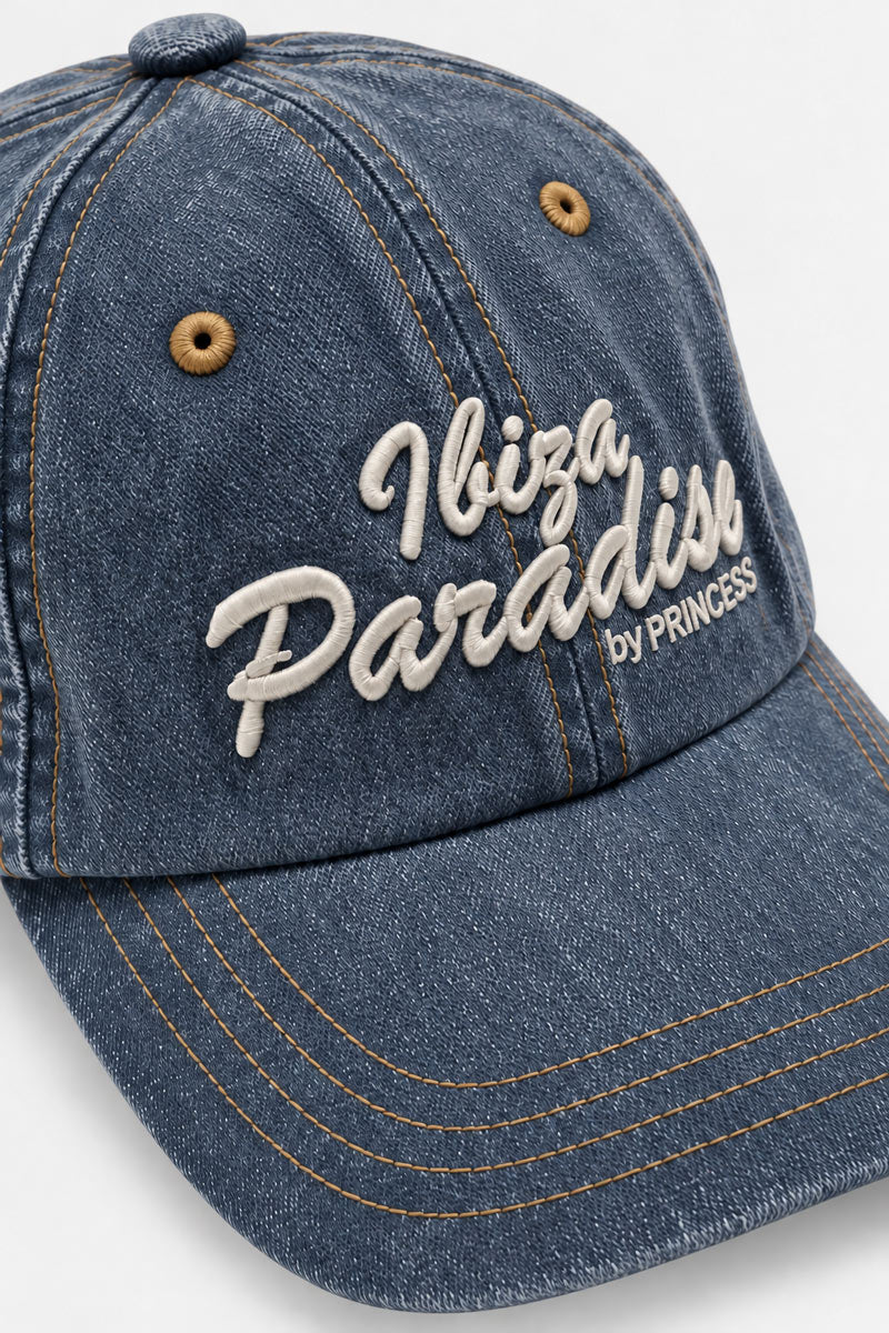Eine Baseballkappe aus hell gewaschenem Denim mit kupferfarbenen Nähten und metallischen Ösen, verziert mit dem weißen Sticktext Ibiza Paradise by PRINCESS auf diesem Accessoire aus Baumwoll-Denim.