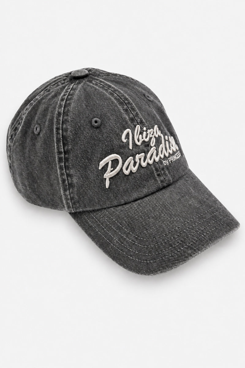 Auf weißem Hintergrund ist eine graue Baseballkappe aus säuregewaschenem Baumwoll-Denim zu sehen. Auf der Vorderseite befindet sich eine weiße Stickerei mit der Aufschrift Ibiza Paradis, darunter steht by PRINCESS.