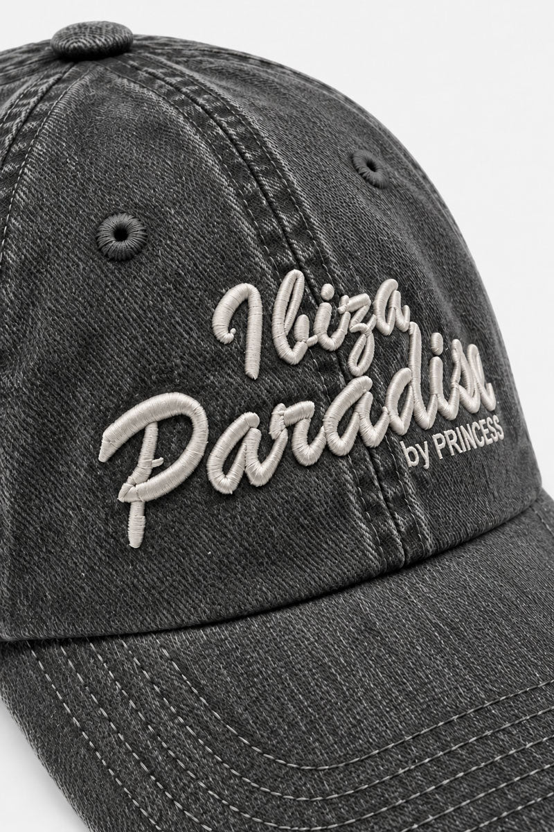 Eine ausgewaschene schwarze Kappe aus Baumwoll-Denim mit abgenutzter Optik, verziert mit einer weißen Stickerei mit der Aufschrift Ibiza Paradise und darunter in kleinerer Schrift by PRINCESS.