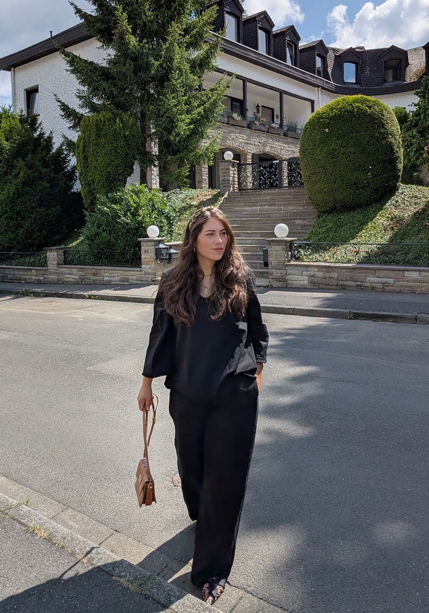 A woman with long brown hair, dressed in a Frogbox by Princess Leinen Bluse mit V-Ausschnitt und langen weiten Ärmeln, walks along a quiet street with a small brown purse. Behind her is a stylish house with manicured bushes and stone steps.
