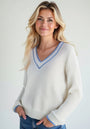 A woman with long, wavy blonde hair smiles gently, wearing a cream ribbed V-neck sweater with blue trim from Princess Goes Hollywood and blue jeans. The light background highlights her and her stylish outfit.