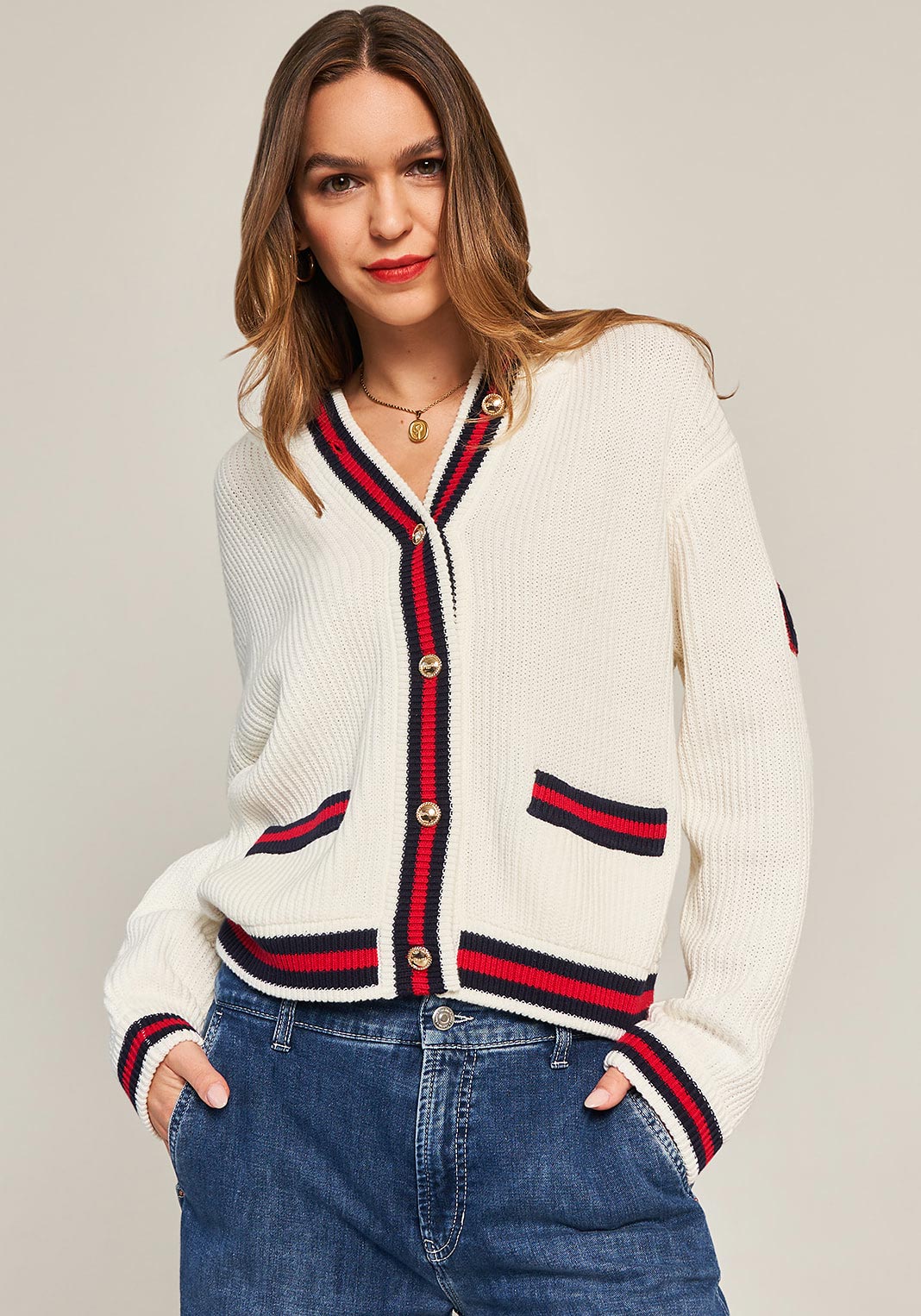 A woman with long brown hair wears a cream-colored Princess Goes Hollywood Strickjacke featuring red and navy trim and Kontrastnähte on the edges and pockets, paired with blue jeans. She stands against a beige background, smiling slightly at the camera.