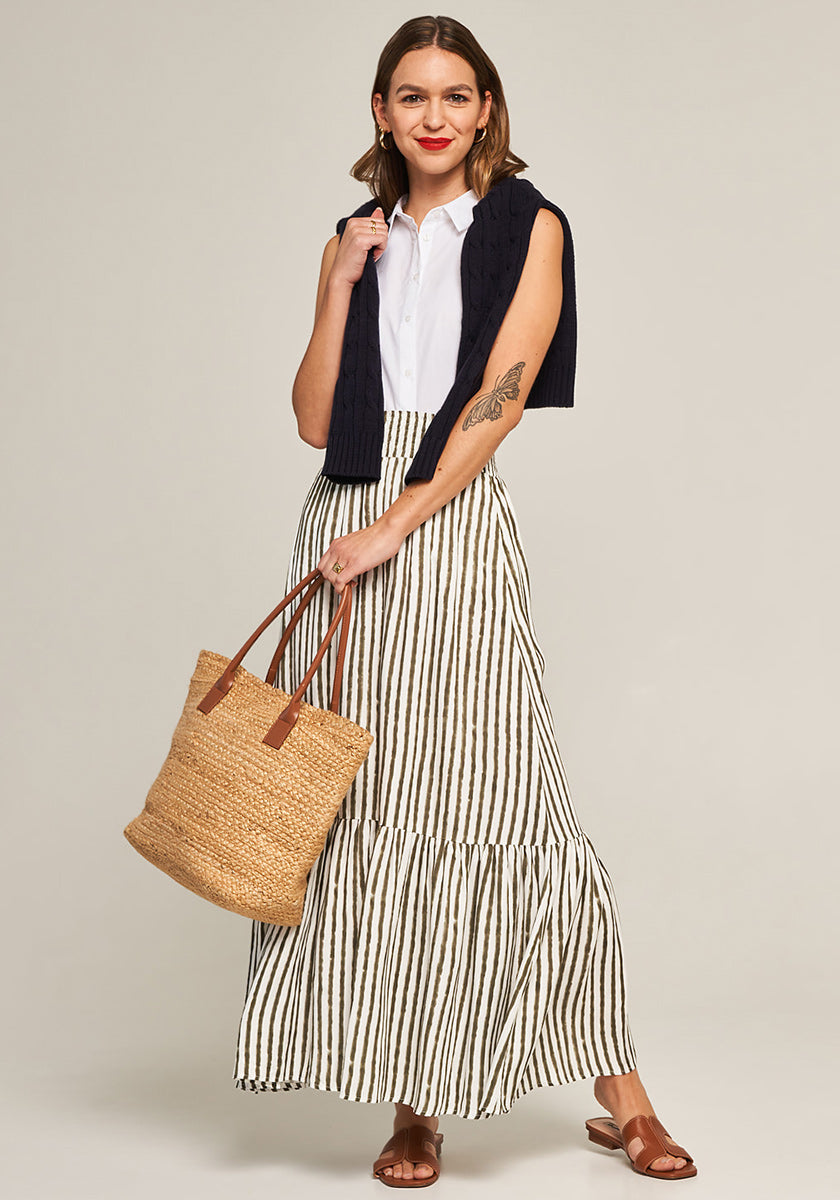 A woman with shoulder-length brown hair and red lipstick stands smiling on the strand, wearing Princess Goes Hollywood’s white button-up shirt, navy sweater over her shoulders, striped long skirt, brown sandals, and holding a large raffiabast tote bag.