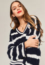 A woman with long brown hair wears a fuzzy navy and white Zopfstrick sweater with V-Ausschnitt from Princess Goes Hollywood. She accessorizes with gold hoop earrings, a necklace, and rings, her red lips poised as she looks down against a neutral backdrop.