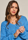 Eine Frau mit welligem braunen Haar trägt einen blauen Zopfstrick-Pullover mit V-Ausschnitt von Princess Goes Hollywood, dazu goldene Uhr, Ringe und Anhängerkette. Sie steht vor neutralem Hintergrund und lächelt selbstbewusst in die Kamera.