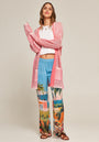 A woman stands against a plain background wearing a pinker Cardigan im Used-Look by Princess Goes Hollywood, a white crop top, blue wide-leg pants with tropical and house prints, and brown sandals. She smiles slightly, arms crossed, radiating relaxed style.