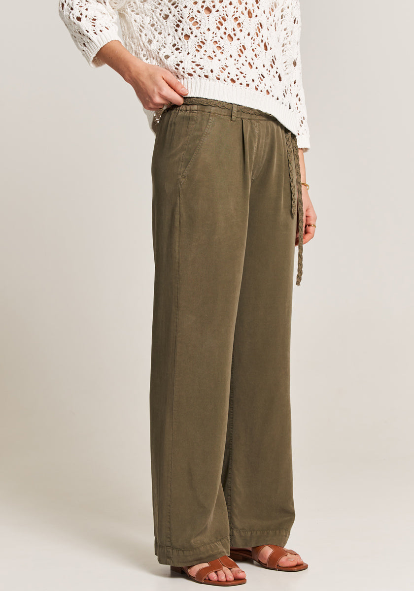 A person stands sideways in a white crochet knit sweater and loose olive-green Lockere Hose with elastic waistband and tie belt from Princess Goes Hollywood, paired with brown strappy sandals, against a plain light background.