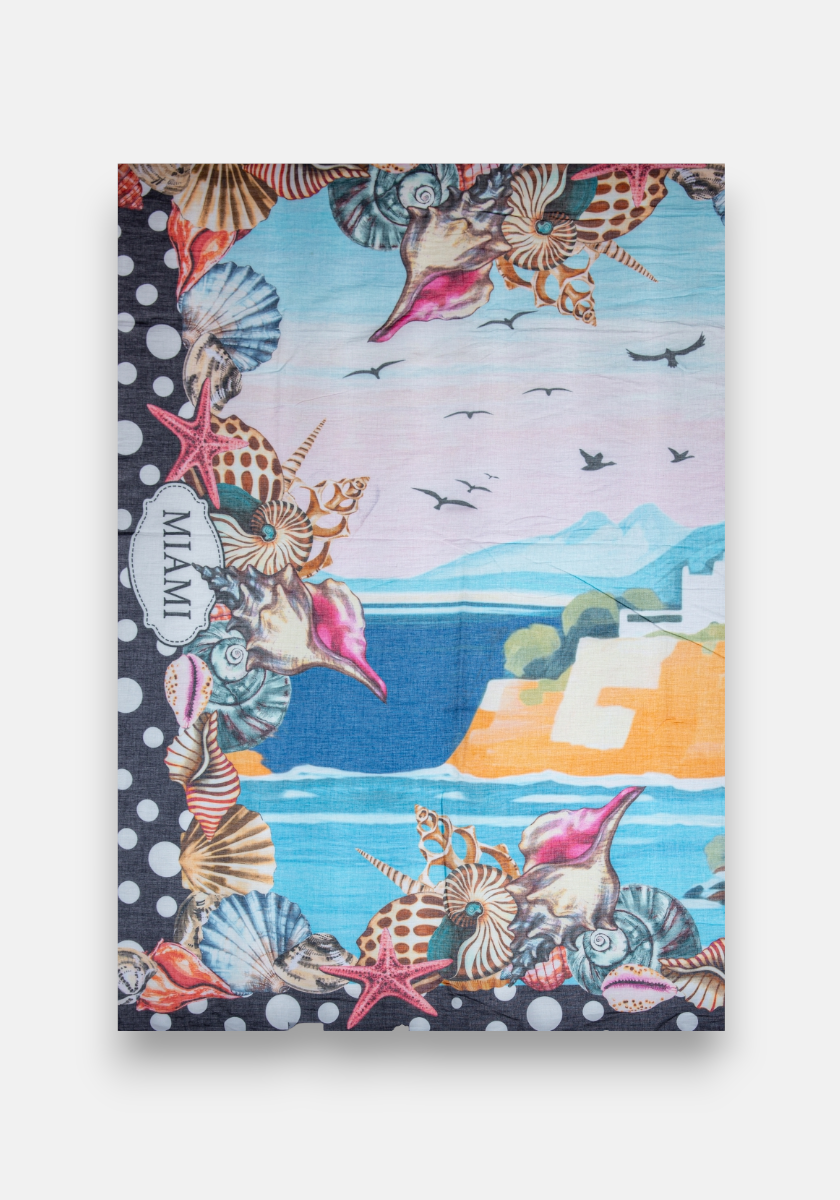 The Pareo Miami by Princess Goes Hollywood is a colorful rectangular wrap featuring blue water, cliffs, flying birds, and white buildings. The border has black and white polka dots, seashells, conch shells, and “MIAMI” in a white oval on the left side.