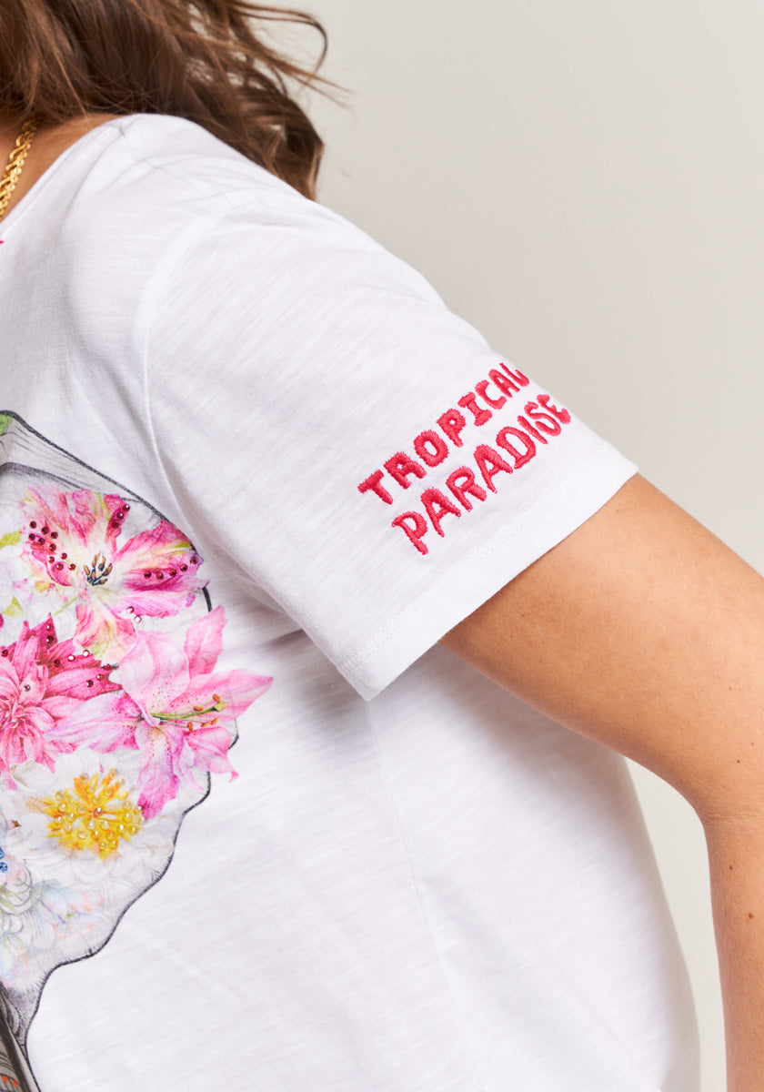A person wears the Princess Goes Hollywood T-Shirt Elefant, a white tee with red embroidered Tropical Paradise on the sleeve, colorful floral designs, and subtle strass detailing on the back. Their arm is slightly bent, hair partly visible.