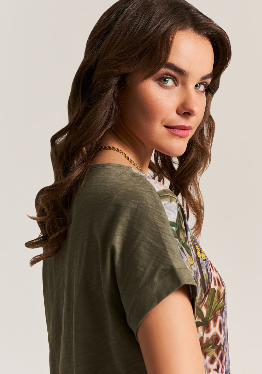 A young woman with wavy brown hair, light skin, and green eyes smiles over her shoulder. She wears a gold chain and an olive-green Princess Goes Hollywood Baumwoll-T-Shirt-Bluse mit Jungle-Print. The background is light beige.