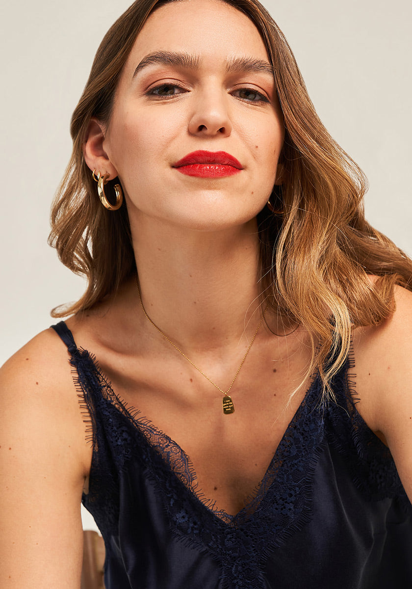 A woman with light skin and long, wavy, light brown hair wears the Seidentop mit Spitze by Princess Goes Hollywood, styled with gold hoop earrings, a delicate pendant necklace, and red lipstick. She gazes confidently at the camera against a plain background.