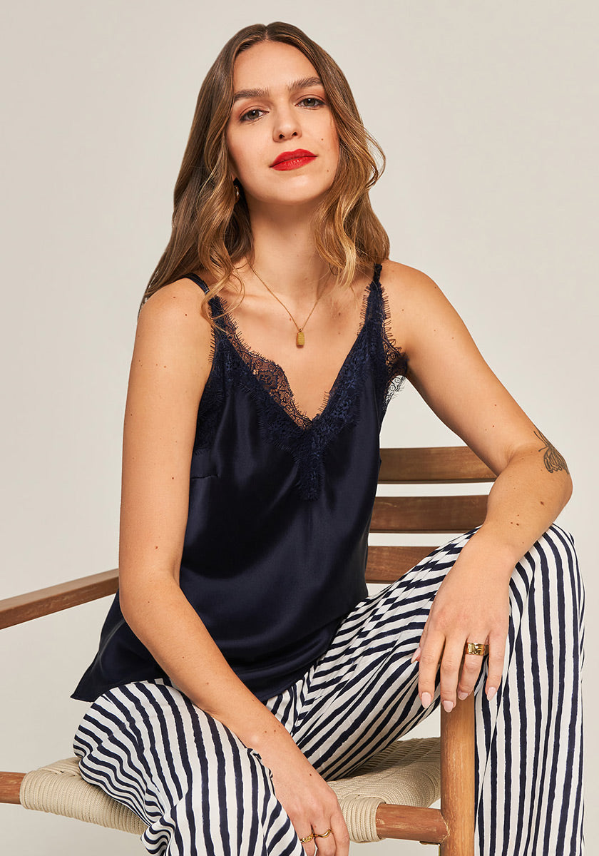 A woman sits on a wooden chair in the Seidentop mit Spitze by Princess Goes Hollywood—a navy camisole with schmale Träger and zarte Spitze trim—paired with black-and-white striped pants, gold jewelry, wavy brown hair, and red lipstick.
