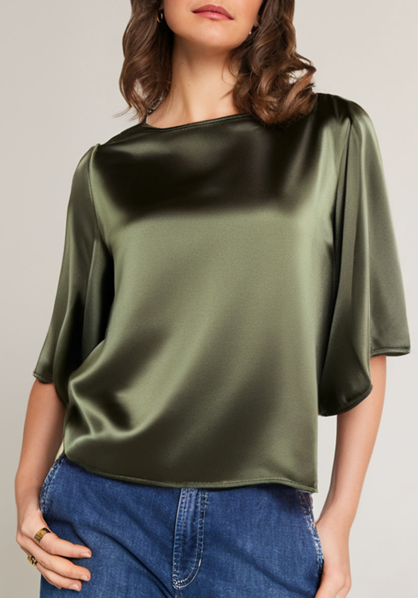 A woman wears an olive green Princess Goes Hollywood Seidenbluse with wide sleeves and a boat neckline, its shiny fabric softly draped. She pairs it with blue jeans, her wavy brown hair partly visible and one hand in her pocket.
