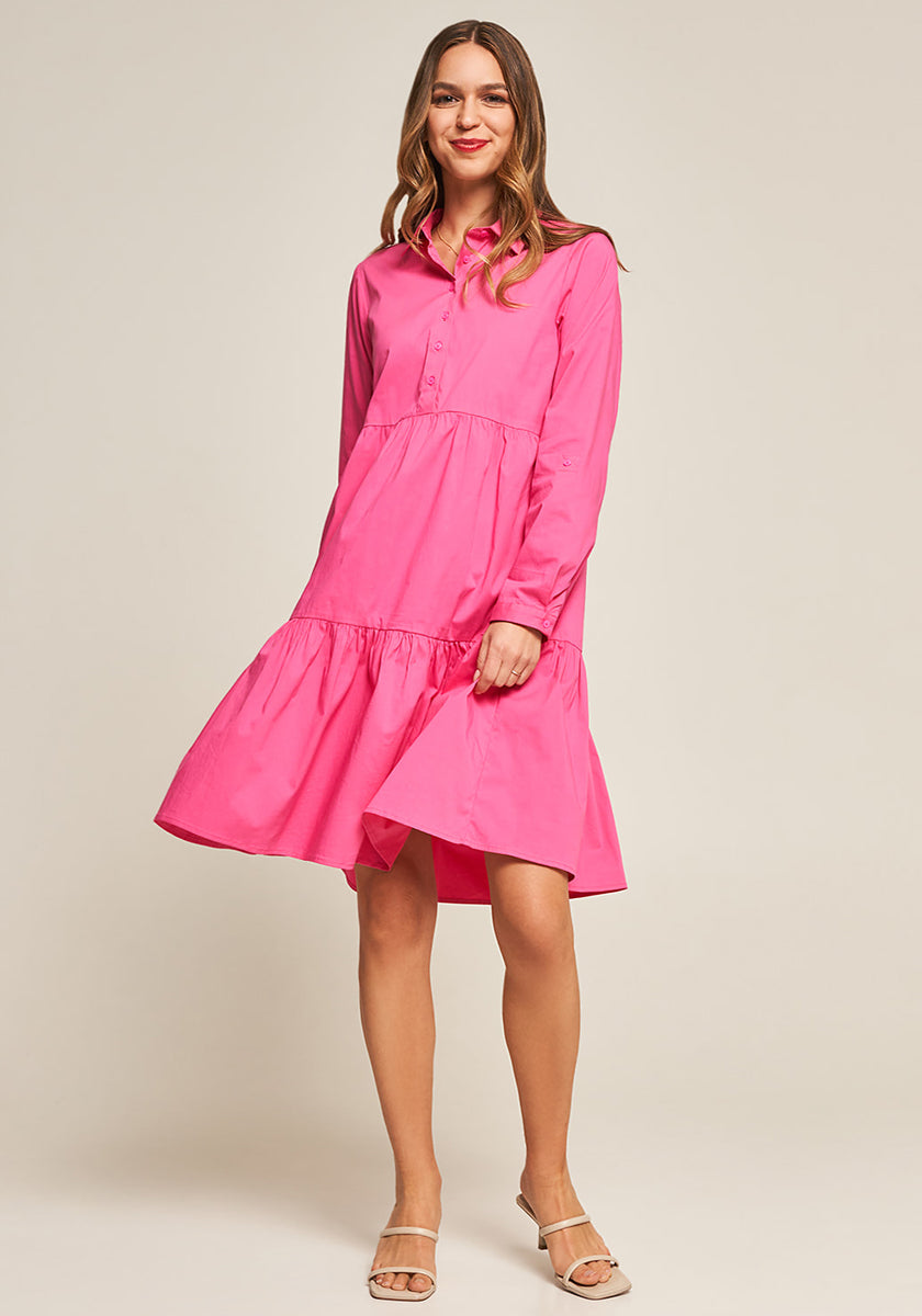 A woman smiles in a bright pink, long-sleeved Kleid aus Baumwollpopeline with elegant Volant details by Princess Goes Hollywood. She has long, wavy hair and beige open-toe heeled sandals. The background is plain and off-white.