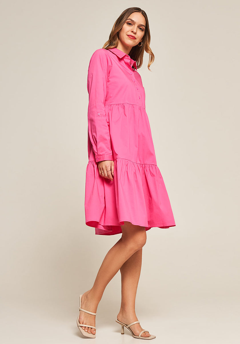 A woman in a bright pink Baumwollpopeline Kleid by Princess Goes Hollywood with Volant details and beige heeled sandals stands against a plain light background, smiling and looking to the side with one leg forward.