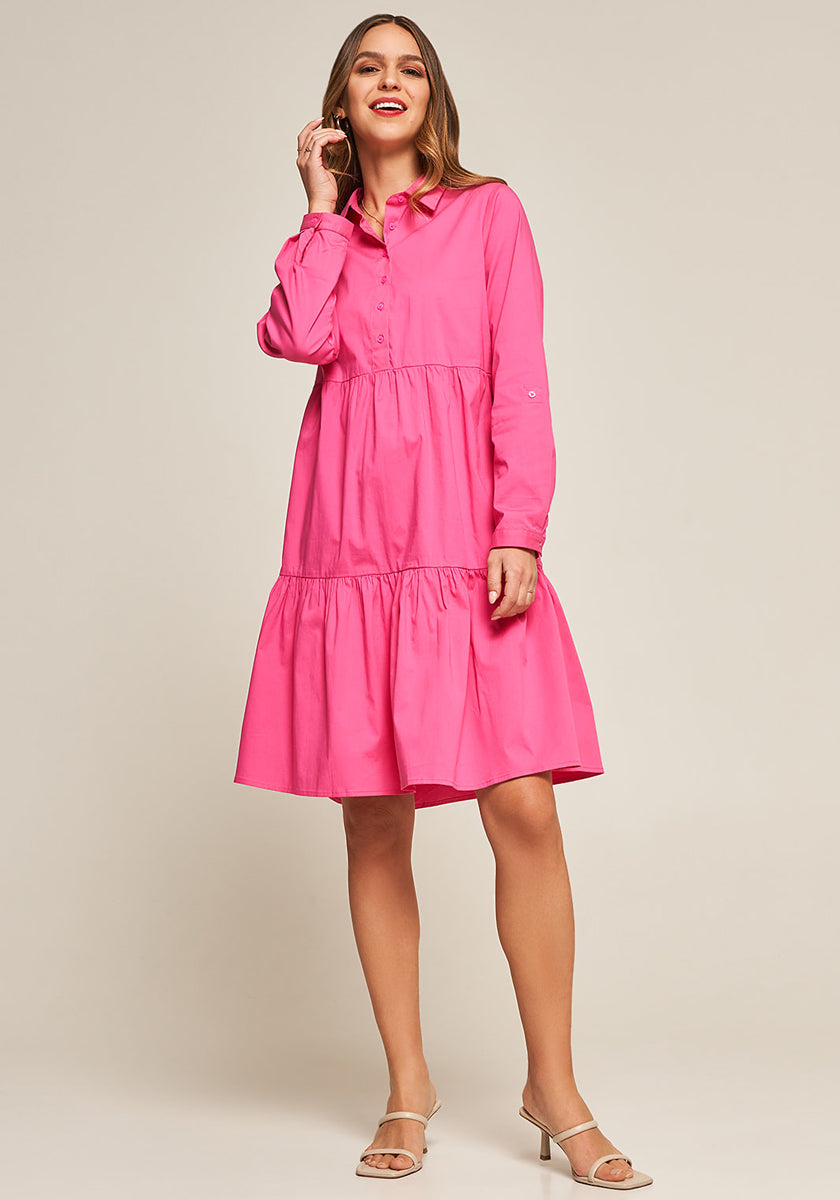 A woman smiles in a bright pink, long-sleeve Volant-Kleid aus Baumwollpopeline by Princess Goes Hollywood that falls to her knees. She has long brown hair, beige heeled sandals, and poses with one hand near her chin against a light background.