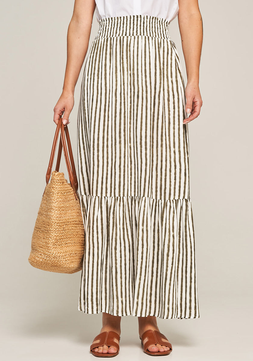 A person faces forward in a white top and a long, high-waisted Princess Goes Hollywood Midirock with brown and white vertical stripes. They carry a woven straw tote and wear brown flat sandals; the background is plain and light-colored.