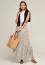 A woman smiles, wearing a white sleeveless blouse, navy sweater over her shoulders, a Princess Goes Hollywood Modal midi skirt with black stripes, brown sandals, and carrying a woven straw tote. A tattoo is visible on her left forearm.