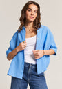 Eine Frau mit welligem braunem Haar trägt eine lockere Leinen-Hemdbluse von Princess Goes Hollywood über einem weißen Tanktop und blauen Jeans. Goldene Accessoires runden den Look ab, während sie vor hellem Hintergrund selbstbewusst nach vorn blickt.