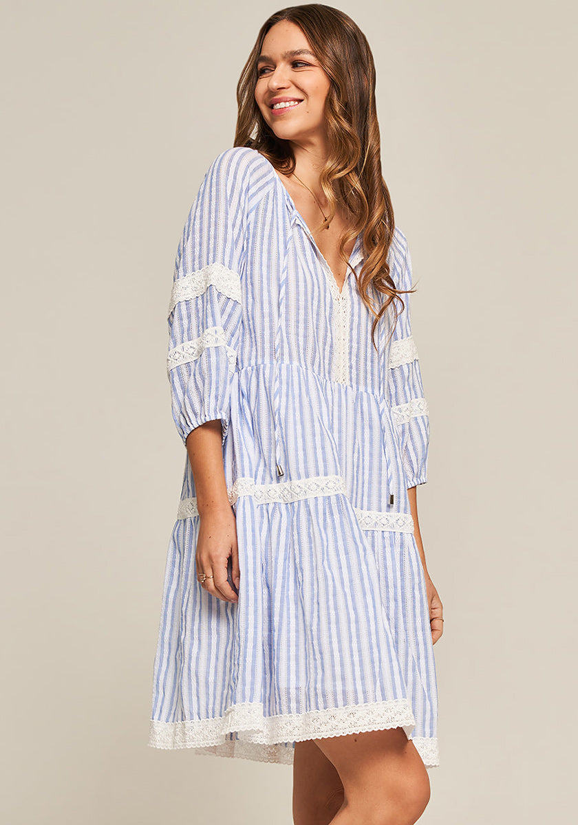 A woman with long wavy hair smiles and looks to the side, wearing a Frogbox by Princess cotton dress with blue and white stripes, lace trim on sleeves, hem, and bodice, plus three-quarter sleeves. The background is plain light beige.