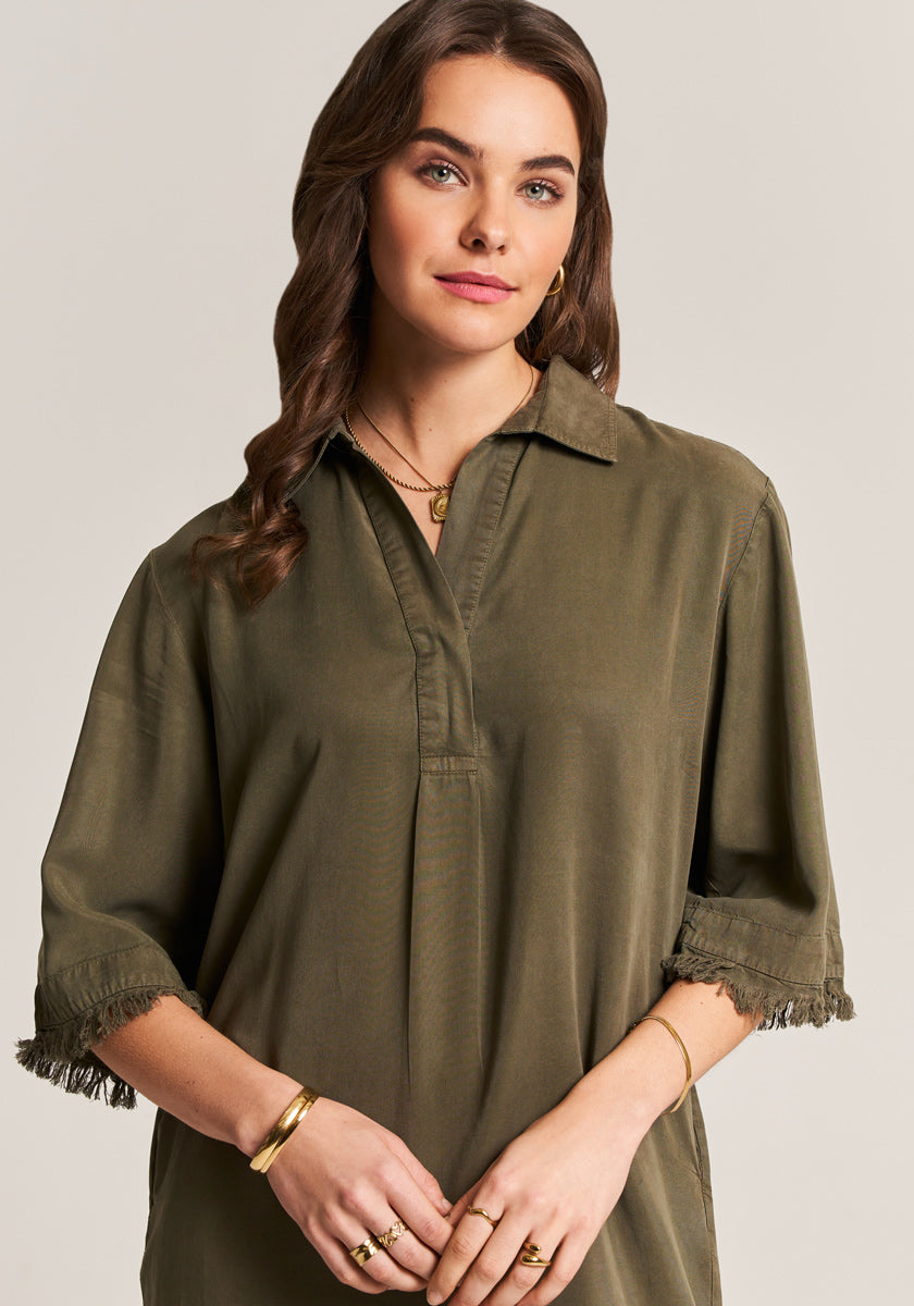 A woman with wavy brown hair wears an olive green Lyocell shirt dress by Princess Goes Hollywood, featuring a Hemdkragen and frayed edges. She accessorizes with gold jewelry and natural makeup, standing against a neutral background.