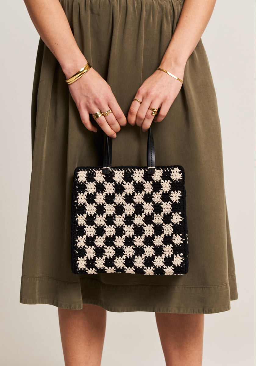 A person wears a knee-length olive green Midirock with elastischer Bund from Princess Goes Hollywood and holds a black and cream crochet handbag with black handles. Their hands are adorned with gold rings and bracelets against a plain background.