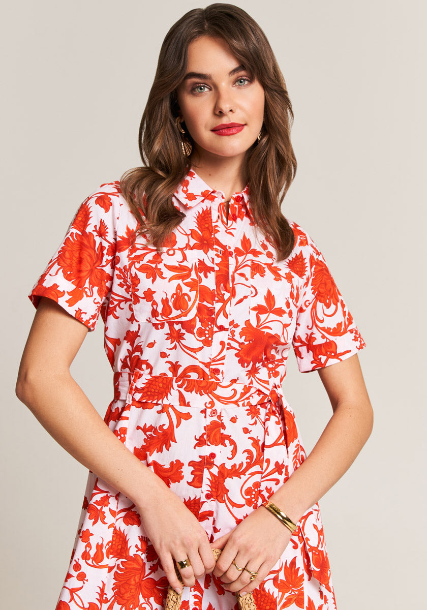 A woman with wavy brown hair models a Princess Goes Hollywood short-sleeved Hemdkragen dress in bold red Blumenprint on white, accessorized with gold hoop earrings and bracelet, smiling softly against a neutral backdrop.