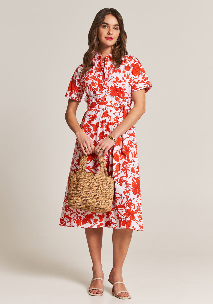 A woman with loose, wavy brown hair poses against a plain background in a white Maxikleid by Princess Goes Hollywood, featuring a bold roten Blumenprint und Hemdkragen. She carries a woven straw handbag and wears white sandals, looking relaxed and pleasant.
