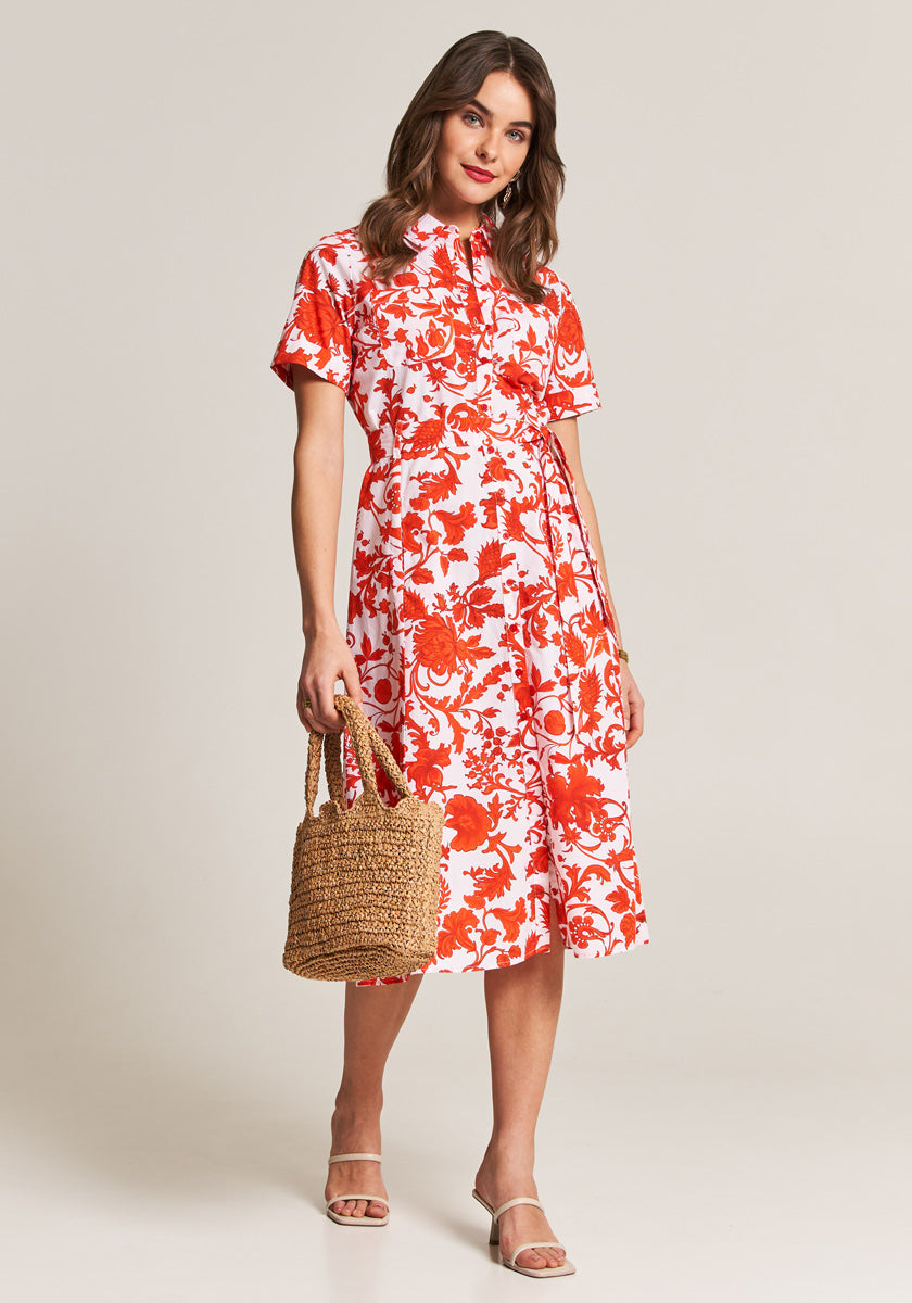 A woman wears a rot-weißes Maxikleid mit Blumenprint und Hemdkragen von Princess Goes Hollywood. Sie hält eine Strohtasche, trägt helle Sandalen, ihr braunes Haar ist offen, und sie lächelt leicht vor neutralem Hintergrund.