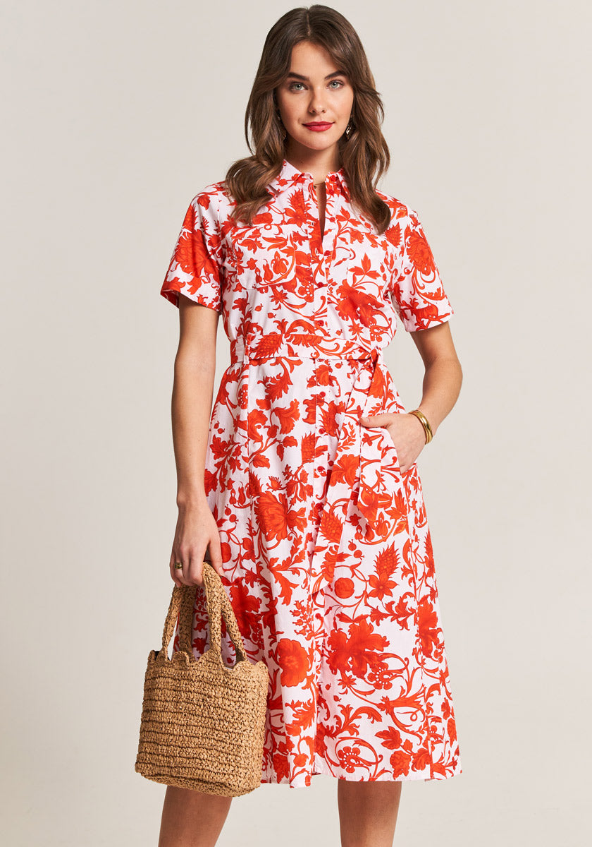 A woman with wavy brown hair models a knee-length, short-sleeved Prinsess Goes Hollywood Hemdkragen dress with a red and white floral print. She stands facing forward, hand in her pocket, holding a straw handbag and smiling softly.