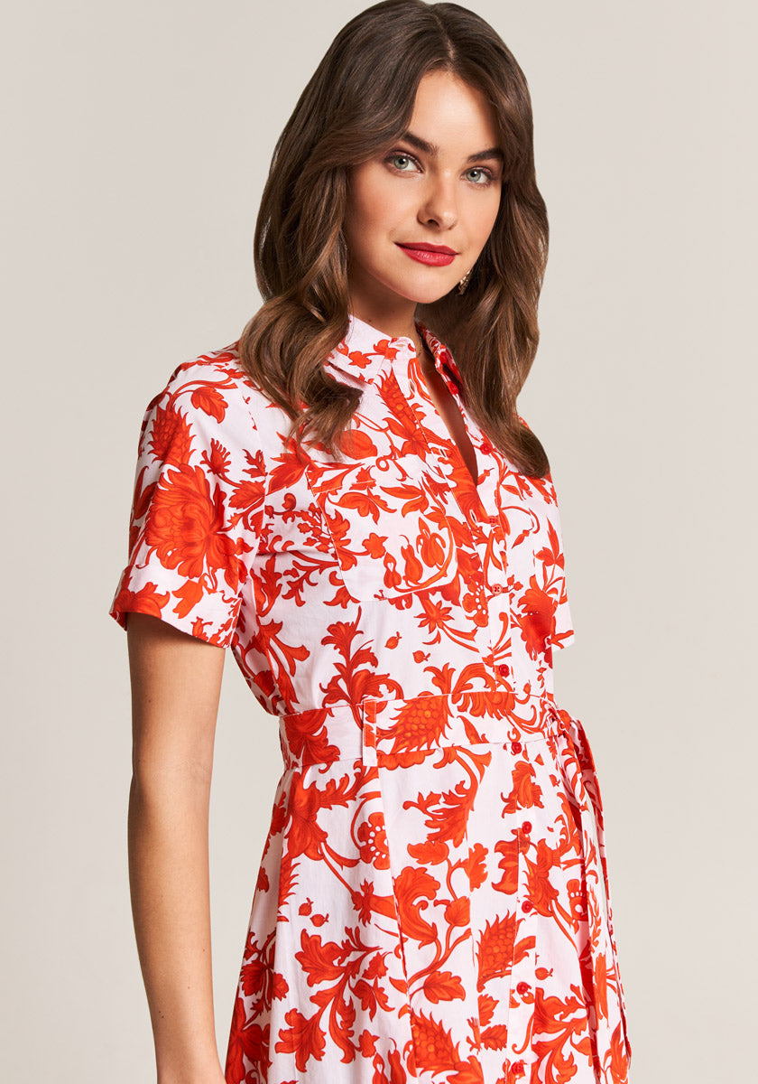 A woman with wavy brown hair and red lipstick wears a Princess Goes Hollywood white maxi dress with red floral print, short sleeves, shirt collar, and front buttons. She stands against a light beige background, smiling softly.