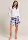 A woman with long brown hair stands against a plain background wearing a white V-neck cable knit pullover and blue and white floral knee-length shorts from Princess Goes Hollywood, paired with beige heeled sandals. Her hands are in her pockets.