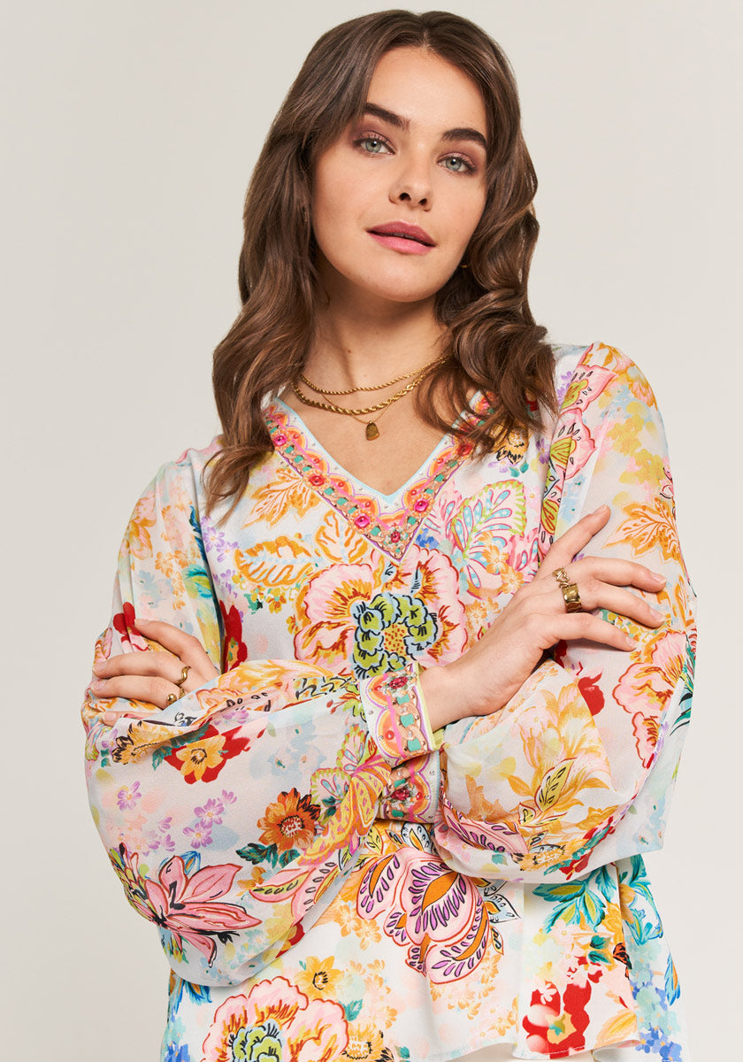 A woman with wavy brown hair wears the Florale Bluse mit V-Ausschnitt by Princess Goes Hollywood, featuring long sleeves and pink-trimmed neckline. She poses arms crossed, looking upward, accessorized with gold jewelry against a light background.