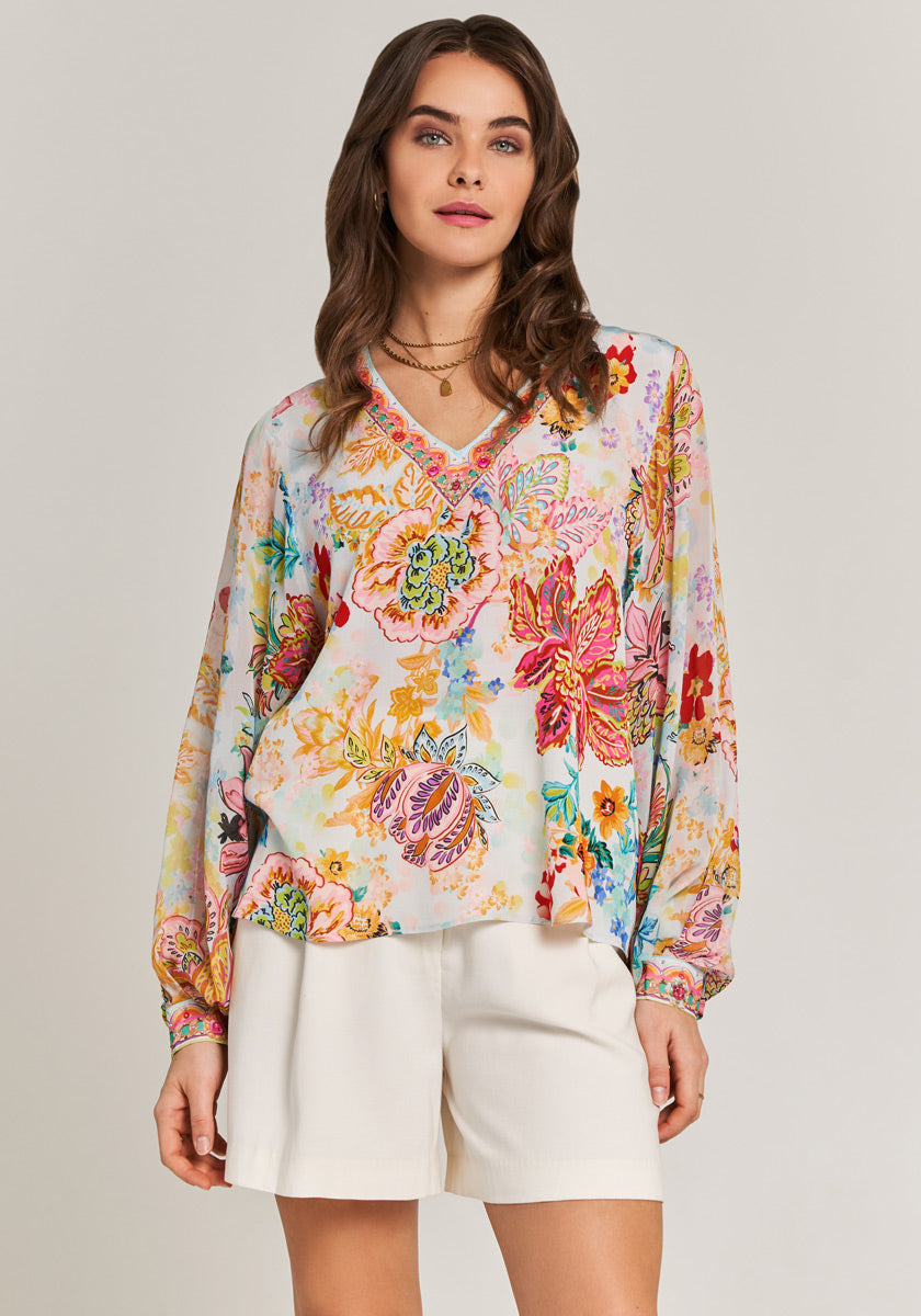 A woman with wavy brown hair wears the Florale Bluse mit V-Ausschnitt by Princess Goes Hollywood and white shorts, facing forward and looking slightly above the camera. She completes her look with layered necklaces.