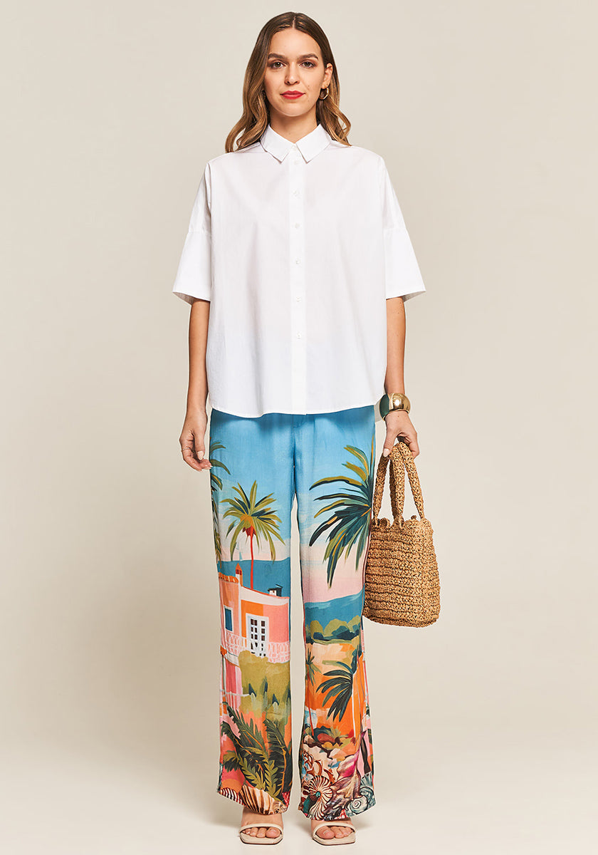 A woman faces forward, wearing a loose white short-sleeve button-up shirt and the Princess Goes Hollywood Hose Miami, featuring a vibrant tropical print. She carries a woven handbag, wears sandals, her wavy hair is loose, and she smiles slightly.