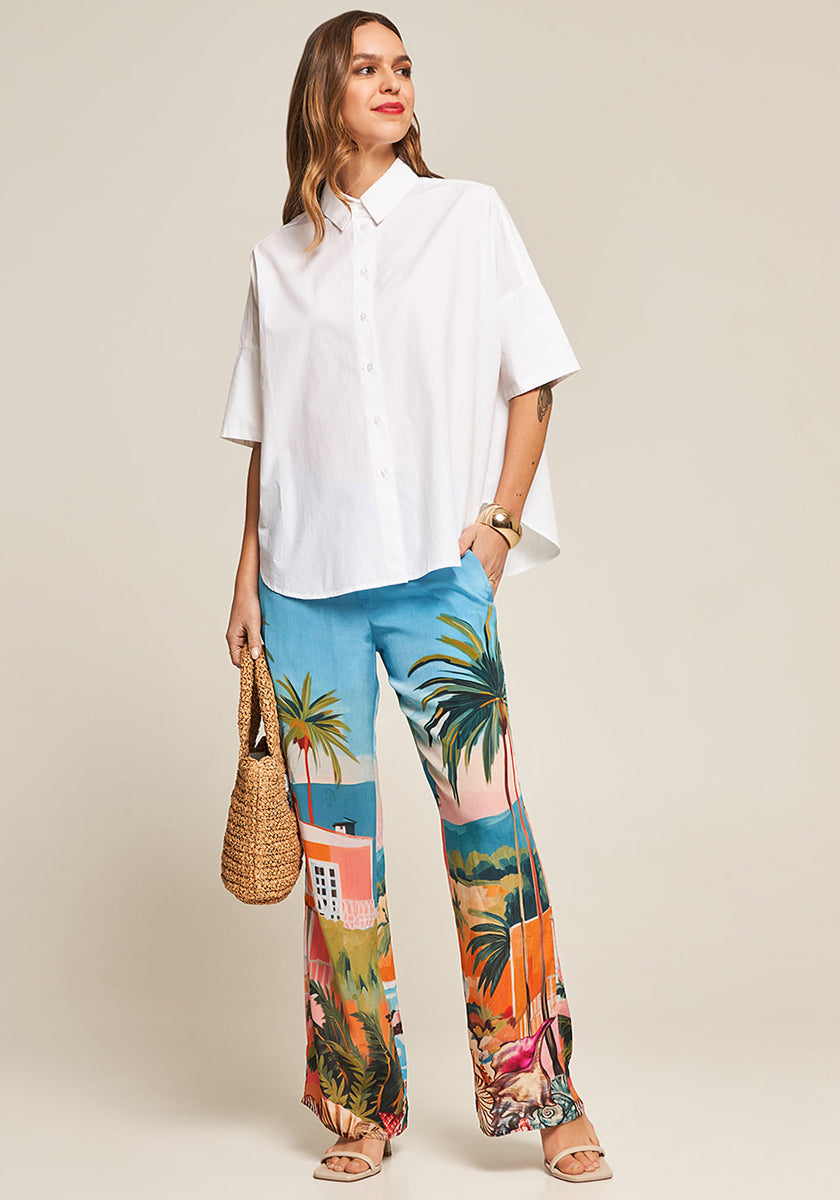 A woman smiles in a loose white button-up shirt and Princess Goes Hollywoods Hose Miami—flowy pants with a colorful Miami print. She accessorizes with a woven handbag, tan sandals, and a gold cuff, all set against a plain beige background.