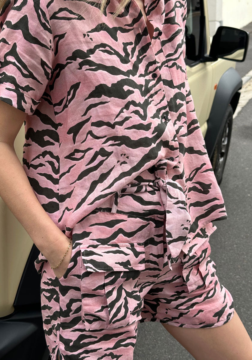 A person in the Princess Goes Hollywood Shorts Tiger mit Gürtelschlaufe, a pink and black tiger-print linen set with pockets and belt loops, stands by a beige vehicle, their patterned outfit partially visible.