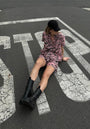 A person in pink and black Princess Goes Hollywood Shorts Tiger mit Gürtelschlaufe, black cap, and boots sits on cracked pavement atop a large white STOP sign, looking down with legs extended and one arm resting behind them.