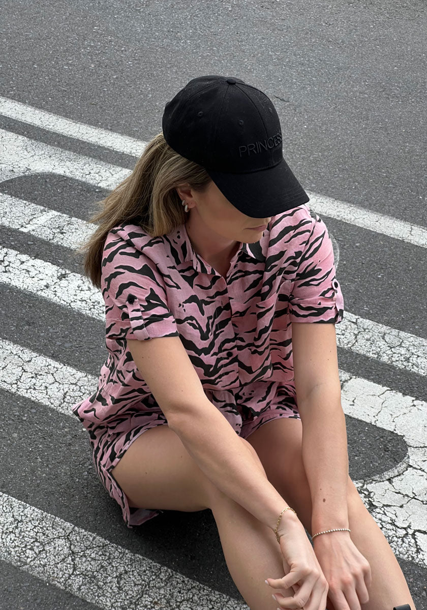 A person with light brown hair, wearing a black cap and Princess Goes Hollywood Shorts Tiger mit Gürtelschlaufe, sits cross-legged on a street with white lines. Their face is mostly hidden by the cap, arms resting on their knees.