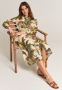 A woman with wavy brown hair sits on a wooden chair, wearing the Princess Goes Hollywood Midi Leinenkleid Safari, featuring a vibrant print of green leaves, flowers, and animals. She rests one arm on the chair; her sandals are flat and brown. Plain light background.