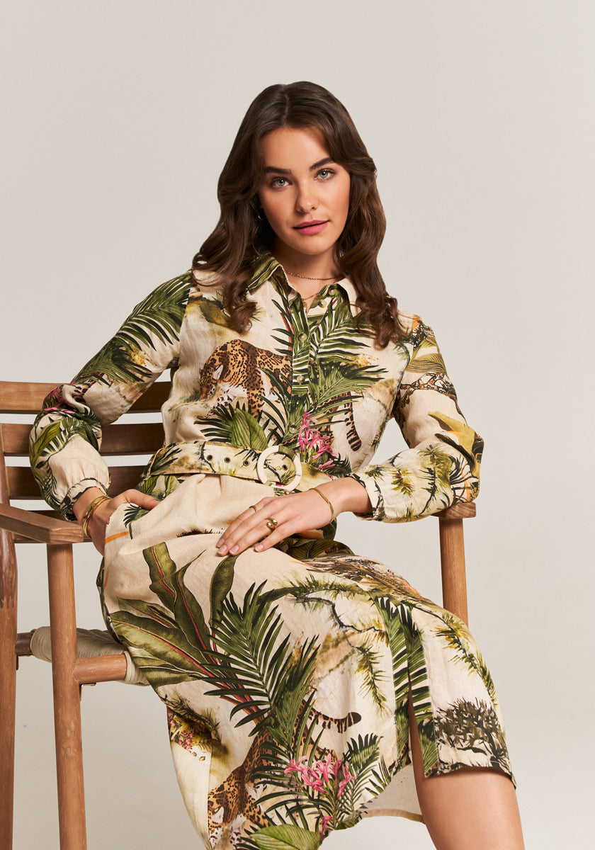 A woman with wavy brown hair wears the Princess Goes Hollywood Midi Leinenkleid Safari, featuring green leaves, pink flowers, and leopards. She sits sideways on a wooden chair, gazing forward with a relaxed expression.