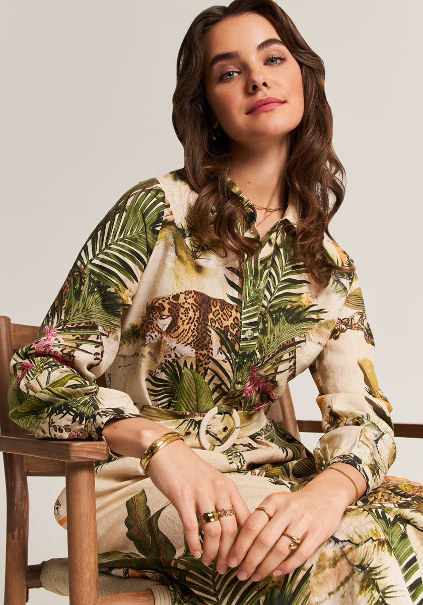 A woman with wavy brown hair sits on a wooden chair, wearing the Midi Leinenkleid Safari by Princess Goes Hollywood, featuring tropical leaves and leopard print. She smiles softly and accessorizes with gold jewelry.