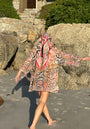 A barefoot person in the Princess Goes Hollywood Tunika Leo-Flower and headscarf walks on a sandy beach near large rocks, arms outstretched. Sunlight creates shadows as stone steps with greenery rise in the background. Their back faces the camera.