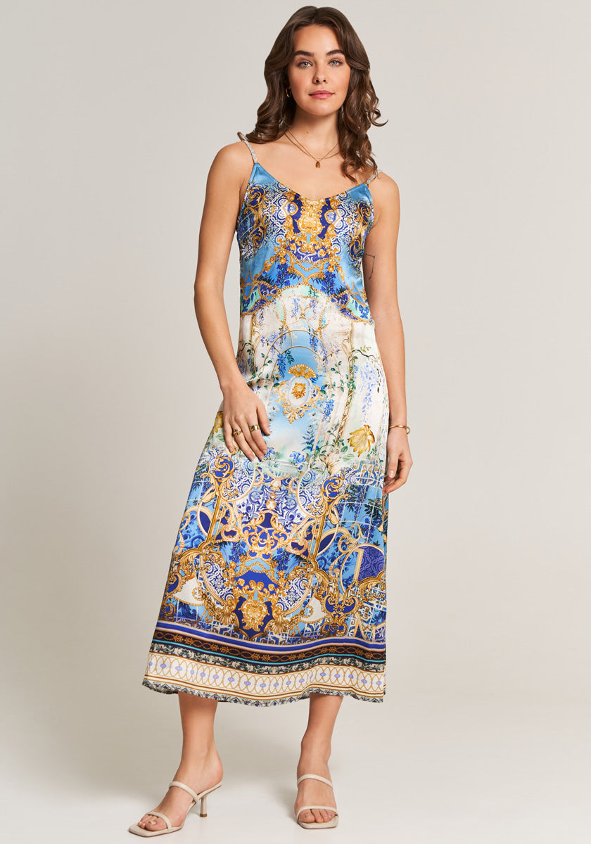 A woman wears a sleeveless, ankle-length Princess Goes Hollywood Seidenkleid with thin straps and an ornate blue, gold, and white baroque pattern. She has wavy brown hair, nude heeled sandals, and delicate jewelry against a plain background.